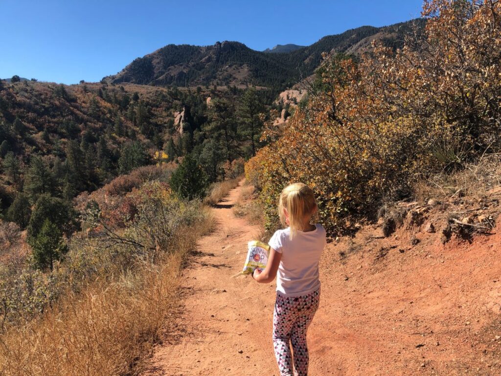 a little girl with a hiking snack on a red dirt trail holding a bag of Skinny Pop popcorn