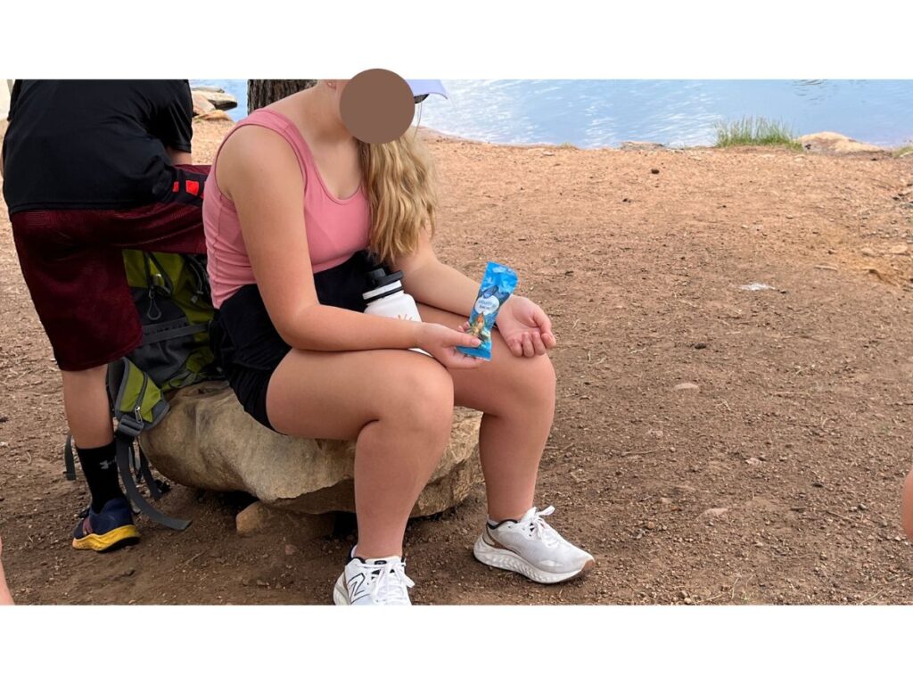 a girl in a salmon colored tank top sits on a rock eating a hiking snack of blue bag of trail mix