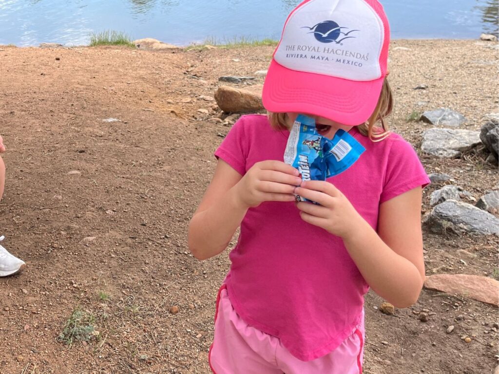 a girl in hot pink with a pink and white hat eats a Cliff Z Protein bar standing up on a dirt/sand area with a lake in the back