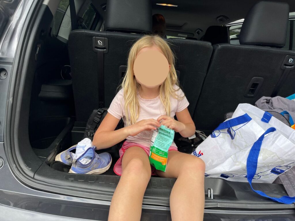 a little girl sits with her legs hanging out of the back hatch of an SUV.  She's holding a bag of yogurt melts after a long hike.