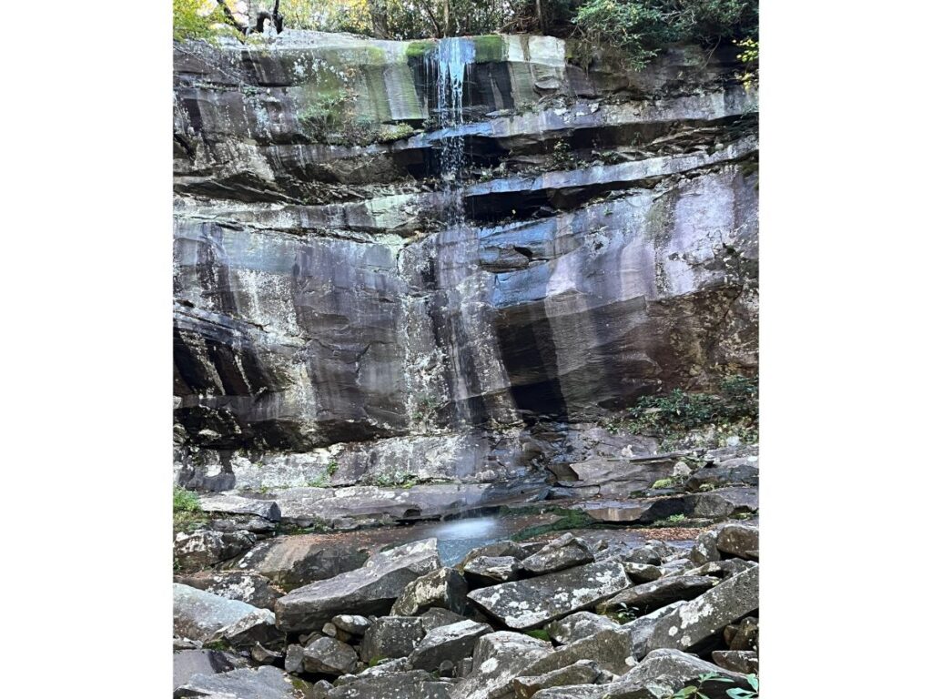 Rainbow Falls in Great Smoky Mountains National Park. A smooth cliff wall with burned sections and a light waterfall coming down. Many large rocks in front