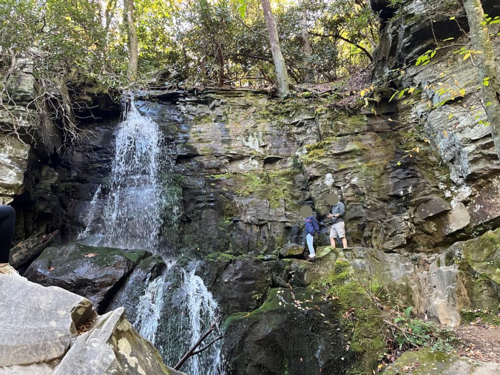 Baskins Creek Falls trail at Great Smoky Mountains National Park leads to a two-tiered waterfall coming down a flat rock, cliffside. Two people stand to the right of the falls higher up. Some of the rock area has green moss