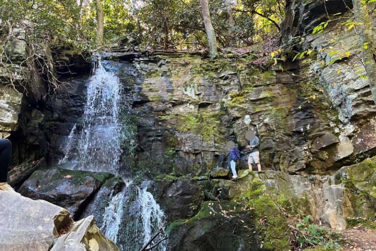 Baskins Creek Falls trail at Great Smoky Mountains National Park leads to a two-tiered waterfall coming down a flat rock, cliffside. Two people stand to the right of the falls higher up. Some of the rock area has green moss