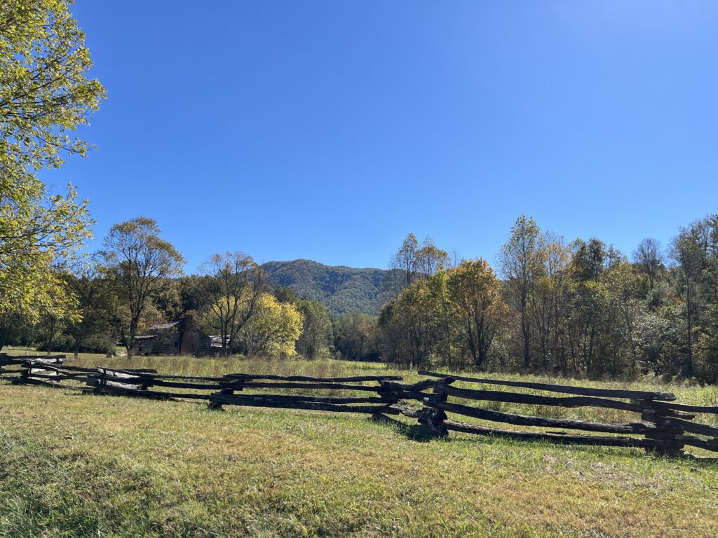 Cades Cove in Great Smoky Mountains National Park 3 day itinerary is a great stop. Looking over an empty, green landscape of grass with trees around the edges and in the distance. A wooden fence in the foreground