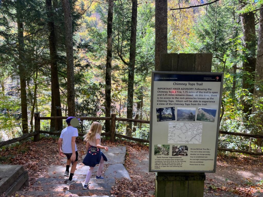 two kids outside, walking past a sign with a notice for the Chimney Tops hiking trail