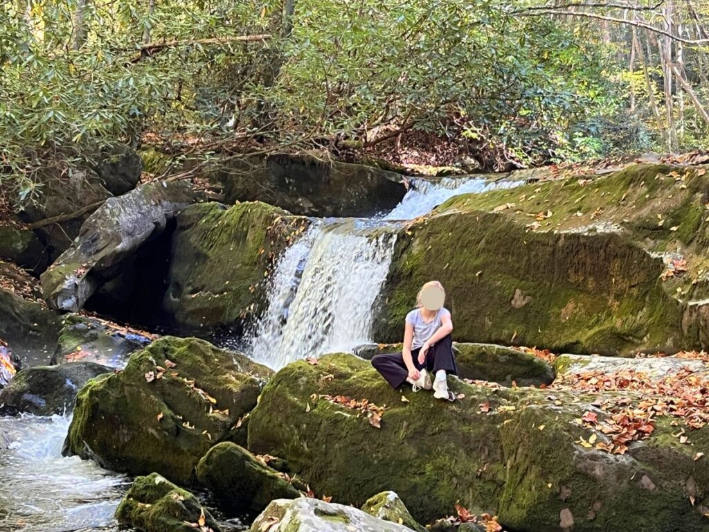 a girl sitting in front of a short waterfall on large rocks on the Lynn Camp Prong trail. This leads to an easy waterfall to see in Great Smoky Mountains National Park