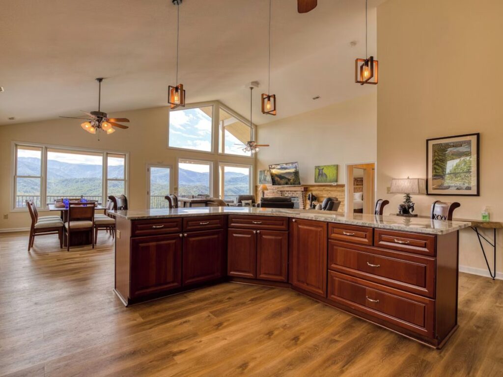 a spacious kitchen with brown cabinets in the island, living area beyond with large windows overlooking distant mountain views