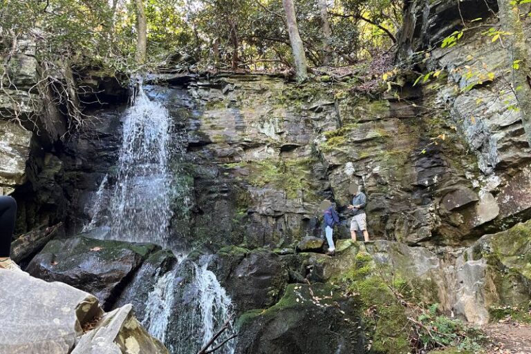 Baskins Creek Falls trail is one of the kid-friendly waterfall trails at Great Smoky Mountains National Park leads to a two-tiered waterfall coming down a flat rock, cliffside. Two people stand to the right of the falls higher up. Some of the rock area has green moss