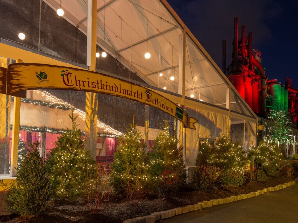 nighttime with the exterior of the Christkindlmarket in Bethlehem, PA.  A yellow Christkindl sign stretches across the endrance, trees below and a white ceiling cover above