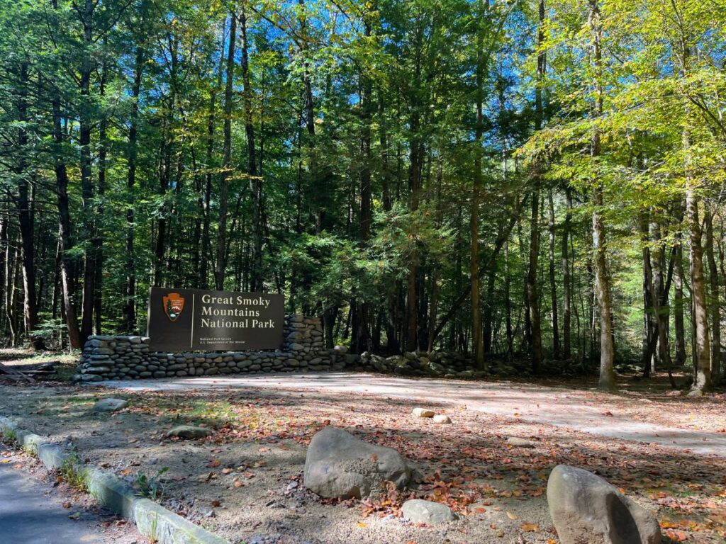 Official park entrance sign for Great Smoky Mountains National Park.  The large brown sign sits low on the ground with lots of green trees around it.  A gravel trail leads to the sign along the side of the road