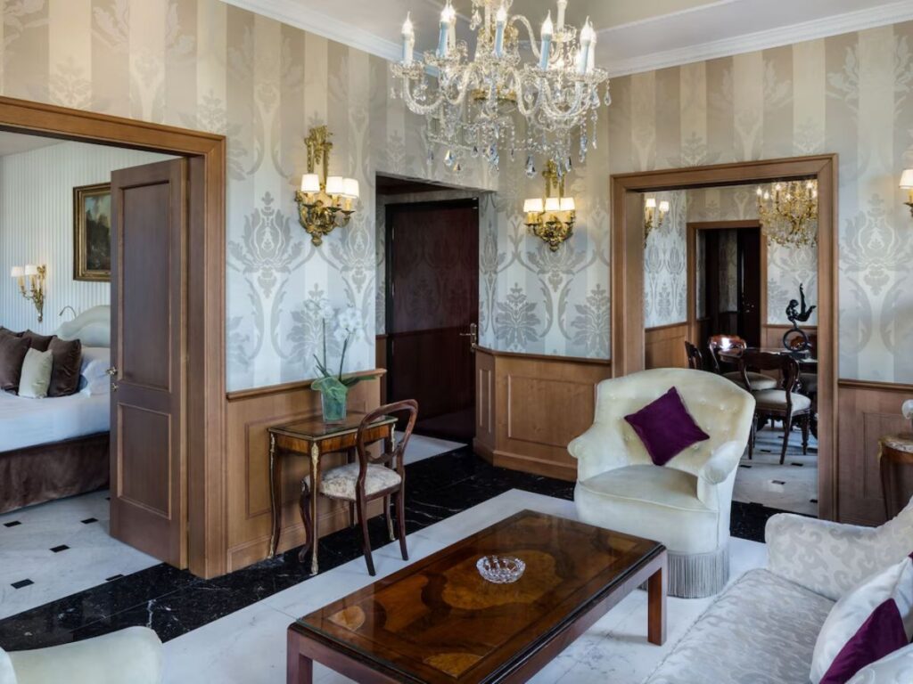 Hotel Baglioni in Rome, Italy.  Looking over an elegant sitting room with a white chair and chandelier.  To the left is a doorway with a bedroom
