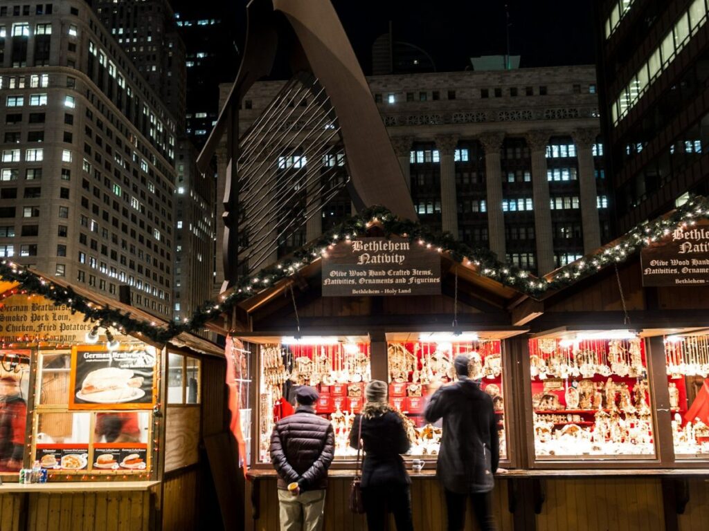 one of the best Christmas Markets in the US is the Chicago market.  Here, 3 people are facing away standing at a lit up wooden hut at nighttime. 