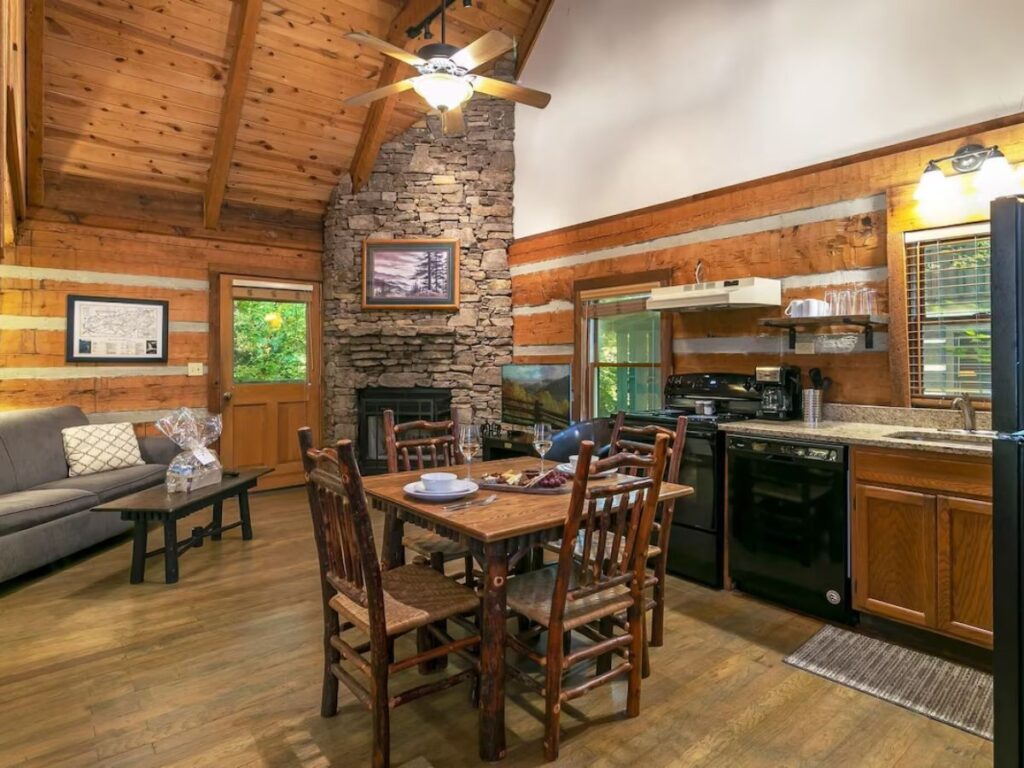inside a large cabin.  Kitchen to the right, 4 person table in the middle, fireplace against the back wall.  Wooden walls and ceilings