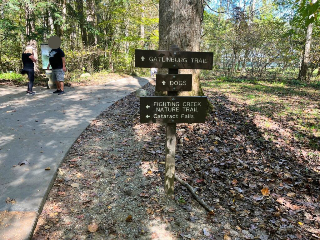 a trail sign pointing to Gatlinburg Trail, Dogs, Fighting Creek Nature Trail, Cataract Falls.  This is one of the easy hikes in Great Smoky Mountains National Park, with a flat trail in the trees.  Two people standing to the left side
