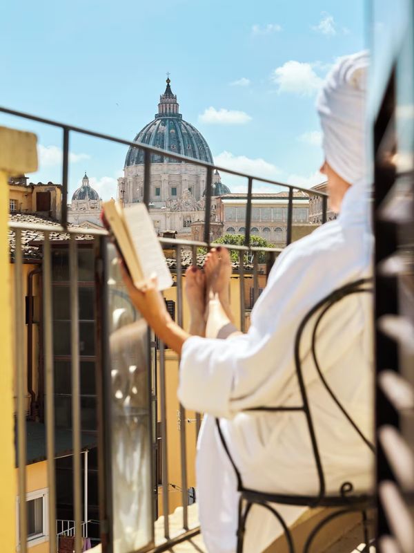 this hotel in Italy shows a woman in a bath robe and hair towel sitting on the balcony with a domed building in the background